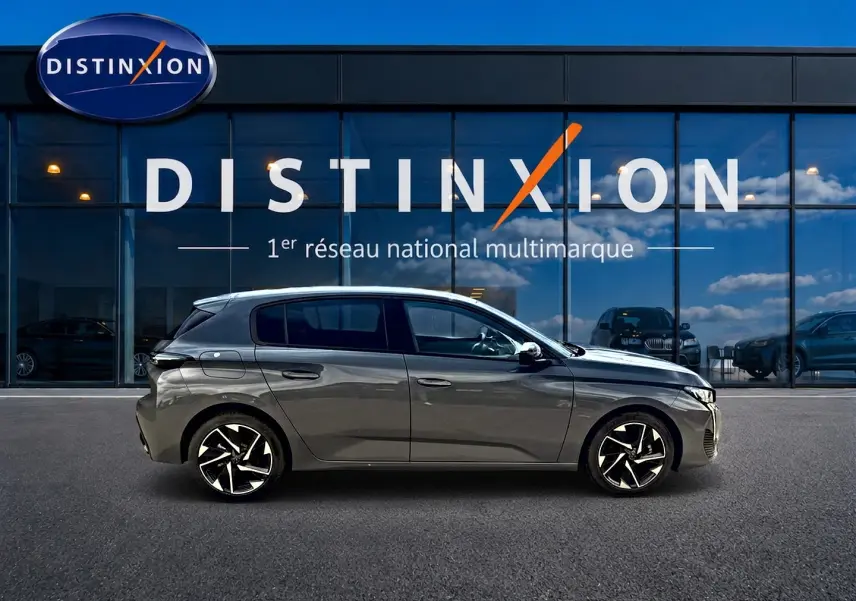 Profil droit d'une Peugeot 308 gris selenium 2025 garée devant une vitrine avec logo Distinxion, jantes alu noires et argentées.