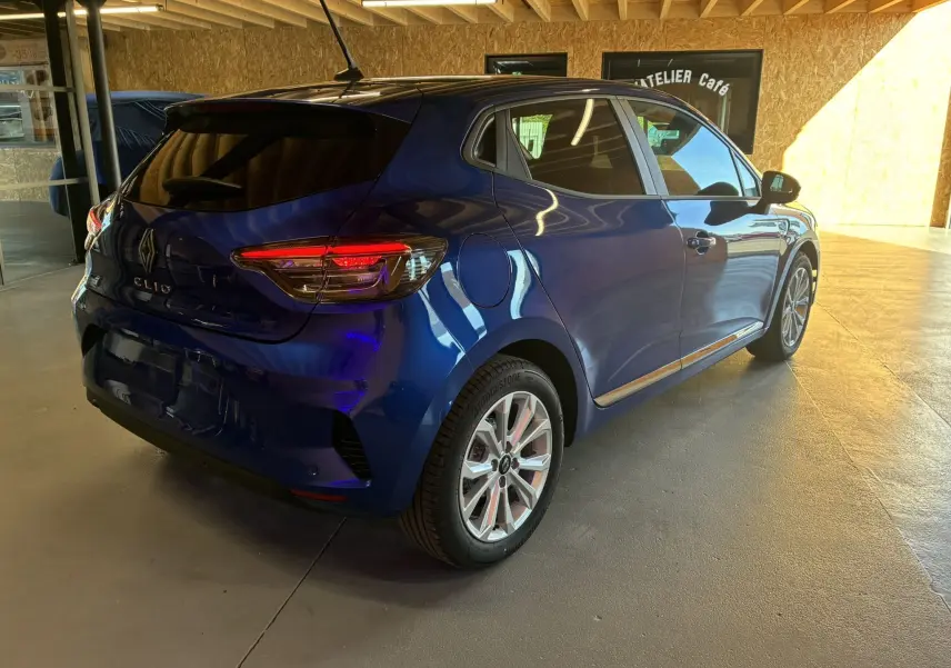 Vue 3/4 arrière droite d'une Renault Clio 2024 bleu iron, avec feux arrière allumés dans un intérieur lumineux.