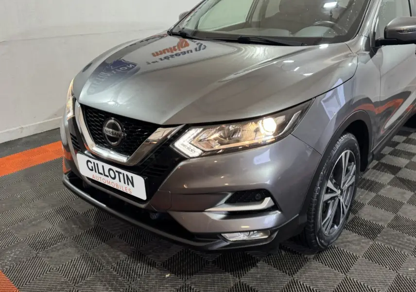 Vue 3/4 avant droit du Nissan Qashqai gris foncé avec phares allumés et calandre chromée distinctive.
