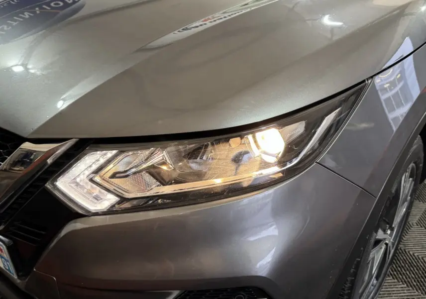 Gros plan sur le phare avant droit allumé d’un Nissan Qashqai gris foncé, avec partie du capot et de la roue visible.