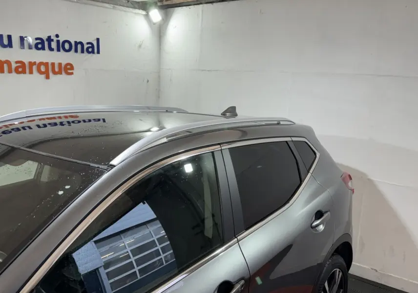 Vue latérale gauche d'un Nissan Qashqai gris foncé avec toit noir et barres de toit aluminium en intérieur showroom.