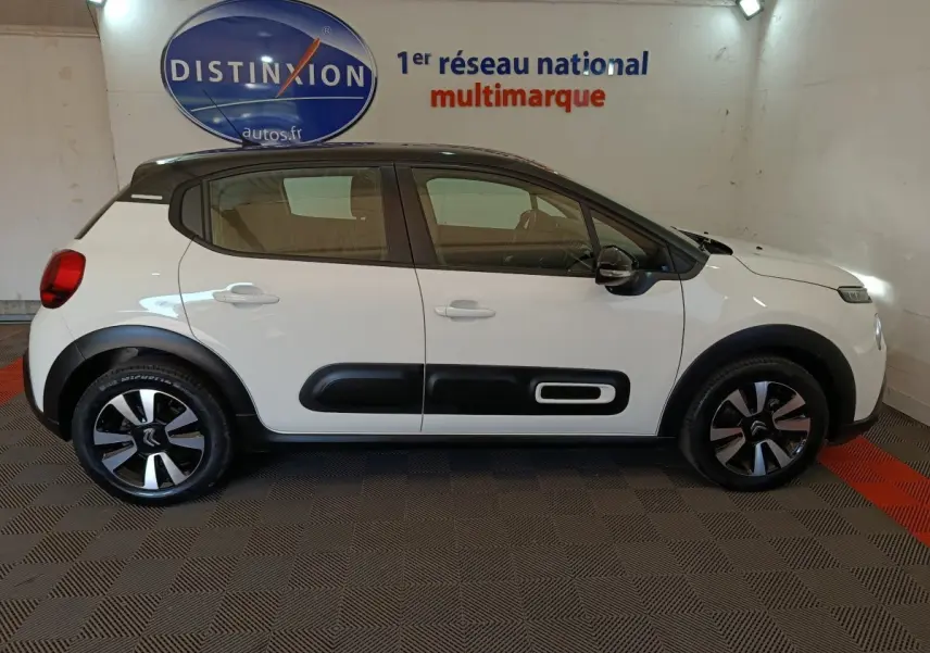 Profil droit d'une Citroën C3 blanche 2021 avec protections latérales noires et jantes bicolores en intérieur showroom.