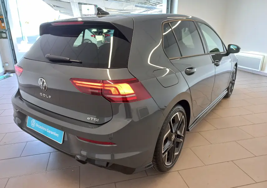 Vue 3/4 arrière droite d'une Volkswagen Golf 1.5 eTSI R-Line gris dauphin métallisé avec feux allumés en intérieur showroom.