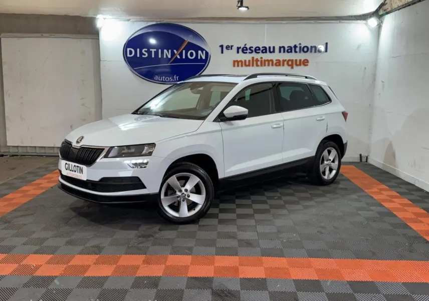 Vue 3/4 avant droite d'un Skoda Karoq blanc stationné en intérieur sur sol noir et orange avec logo Distinxion en arrière-plan.