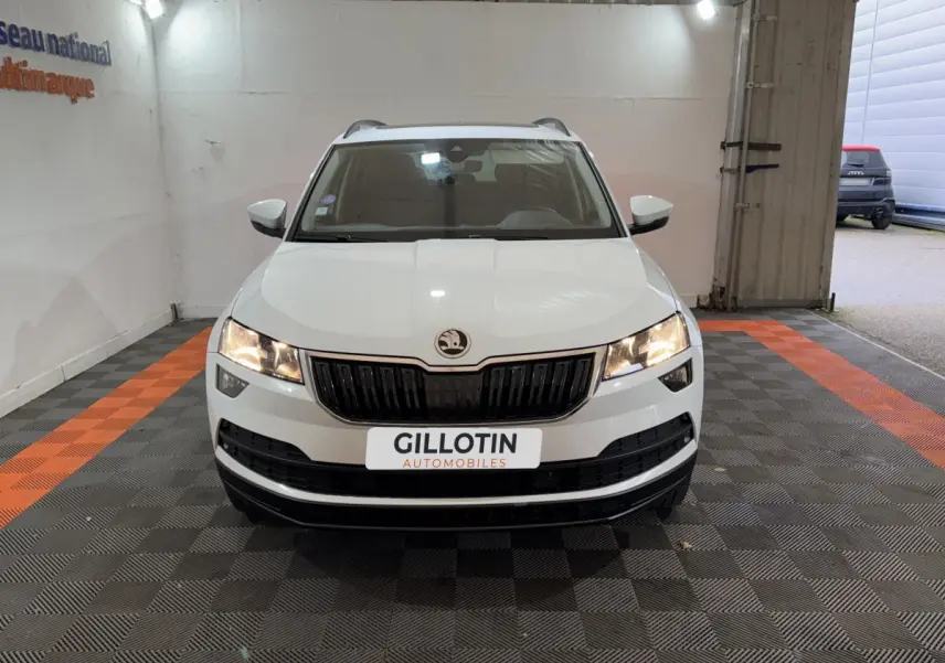 Vue frontale d'un Skoda Karoq blanc avec calandre noire et phares allumés dans un garage éclairé.
