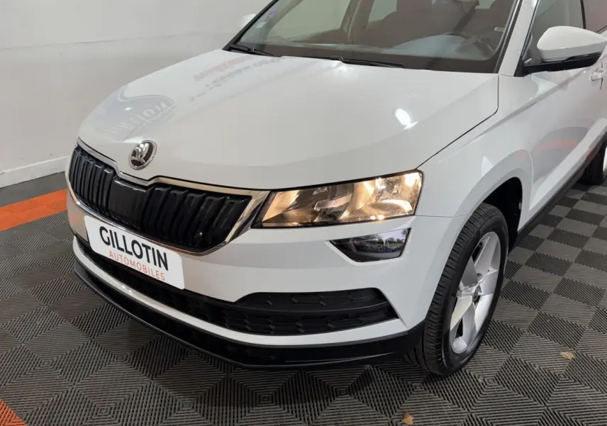 Vue 3/4 avant droite d'un Skoda Karoq blanc avec calandre noire et phares allumés en intérieur showroom.