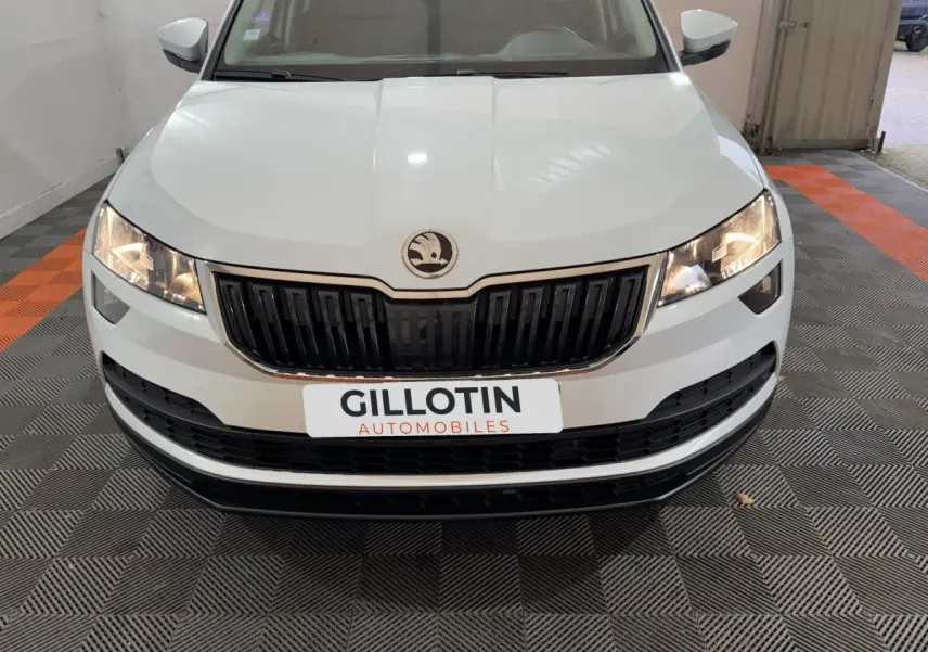 Vue frontale d'un Skoda Karoq blanc avec calandre noire et phares allumés dans un garage.