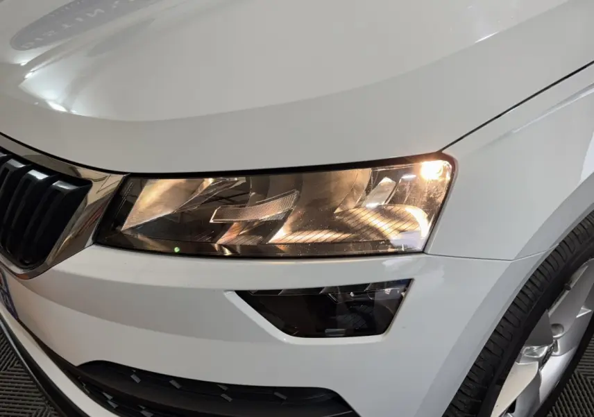 Gros plan sur le phare avant droit allumé d’un Skoda Karoq blanc avec calandre et jante visibles.