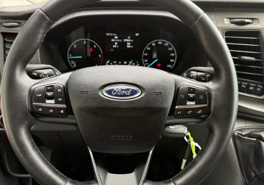Vue rapprochée du volant en cuir noir du Ford Transit Fourgon 2024 avec commandes multifonctions et tableau de bord visible.
