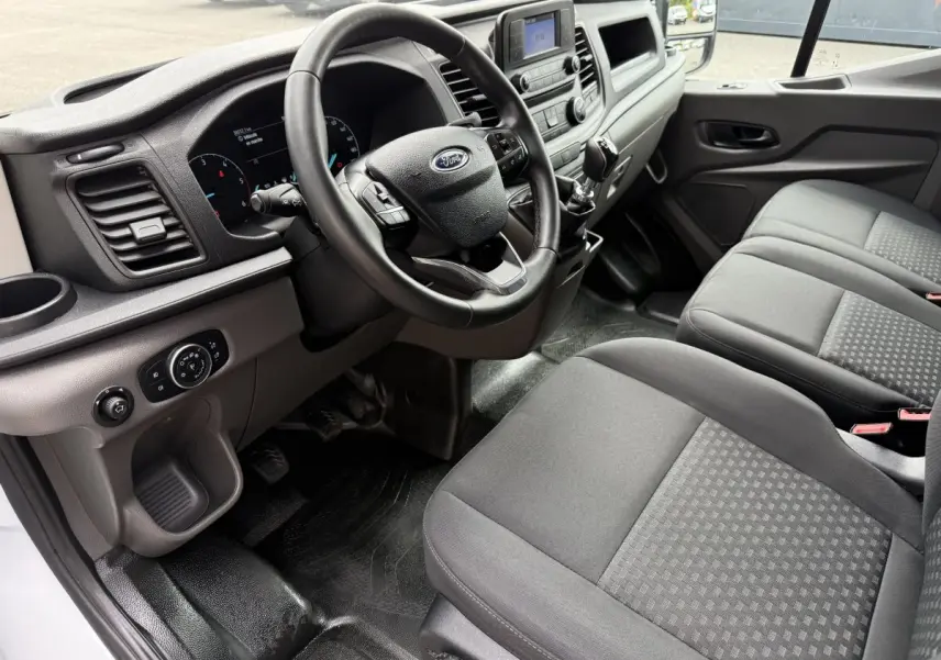 Vue intérieure côté conducteur du Ford Transit Fourgon 2024, montrant le volant cuir, tableau de bord et sièges gris.