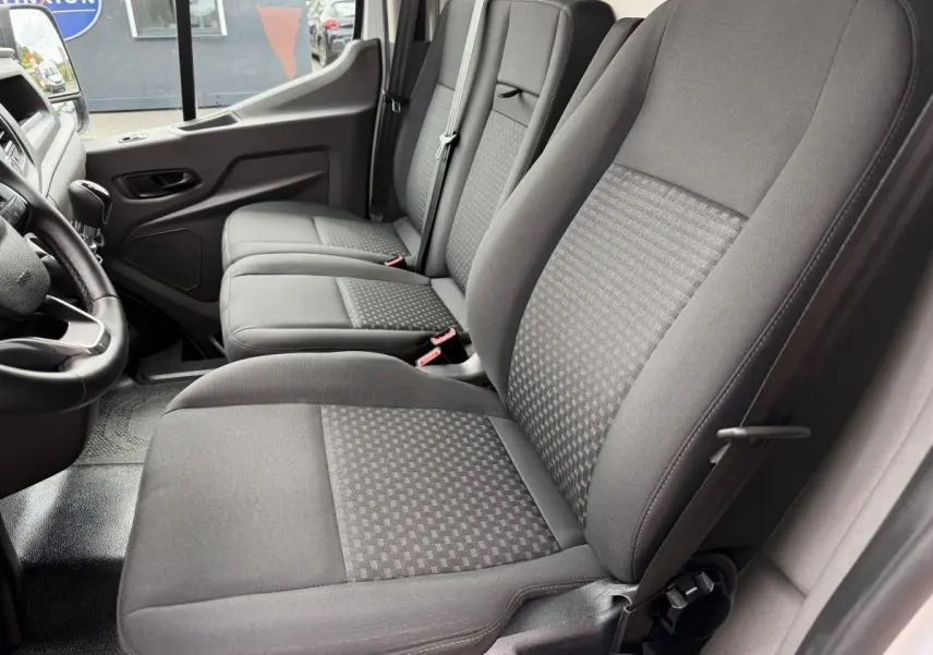 Vue intérieure côté conducteur du Ford Transit Fourgon 2024, montrant les sièges en tissu gris et le volant cuir.