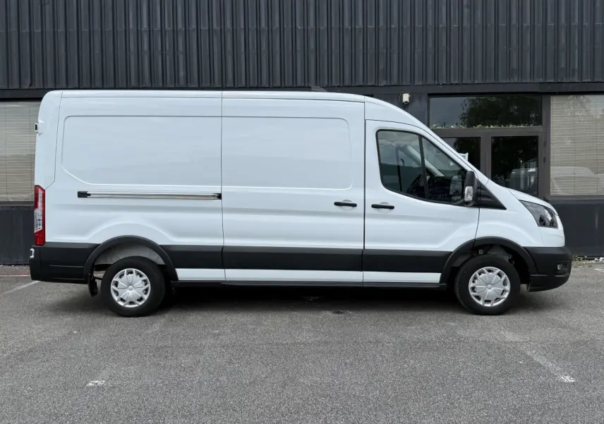 Vue latérale droite d’un fourgon Ford Transit blanc avec bande noire et jantes tôles, stationné devant un bâtiment gris.