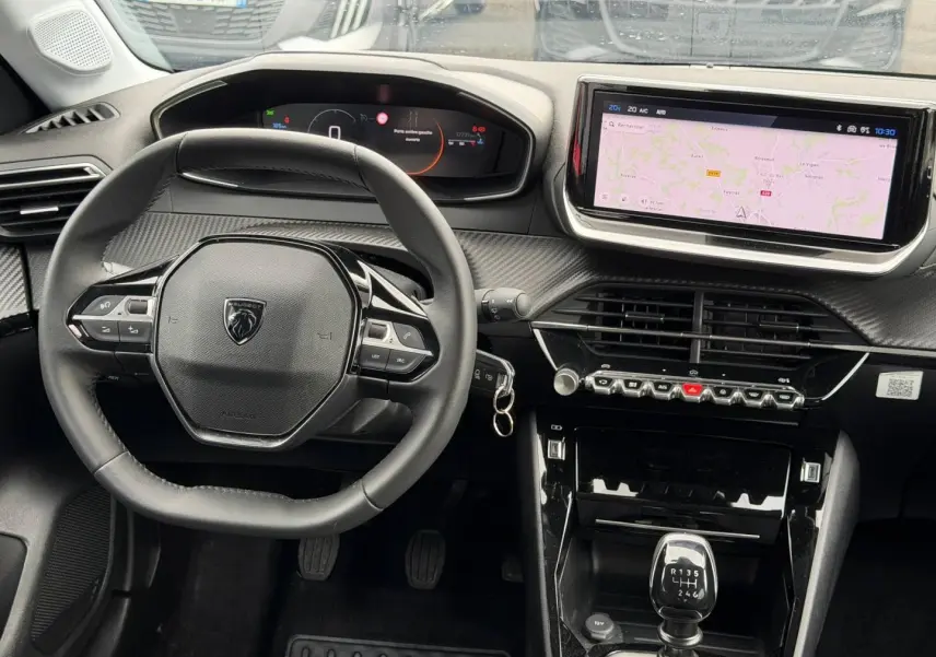 Vue intérieure centrée sur le volant et la console centrale d'une Peugeot 208 noire, avec écran tactile et levier de vitesse manuel.