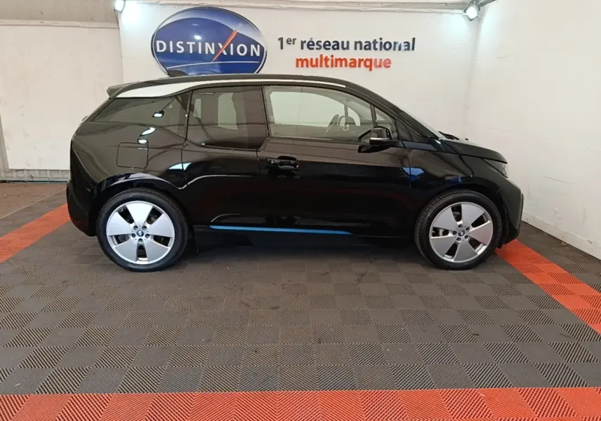 Profil côté gauche d'une BMW i3 120 Ah Atelier noire, compacte et électrique, stationnée en intérieur sur sol gris et rouge.