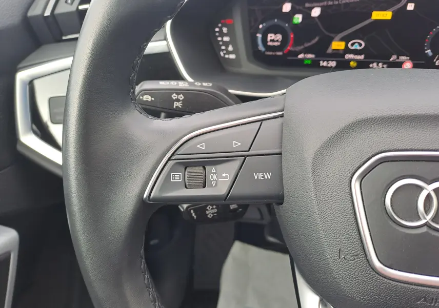 Gros plan sur le volant noir de l'Audi Q3 2023 avec commandes intégrées et tableau de bord numérique en arrière-plan.