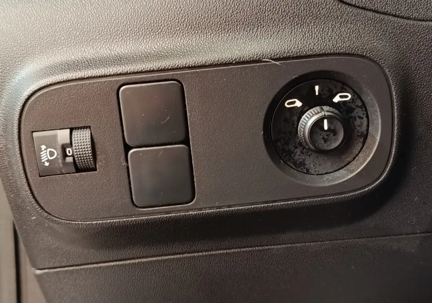 Gros plan sur les commandes noires de réglage des phares et des rétroviseurs d'une Citroën C3 blanche, côté gauche.