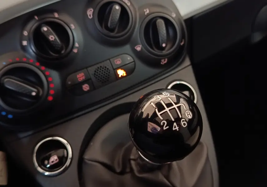 Gros plan sur le levier de vitesses manuel noir et le tableau de bord avec commandes de climatisation d'une Fiat 500 blanche.
