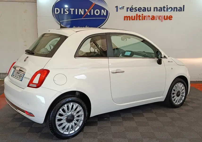 Fiat 500 Hybrid blanc vue 3/4 arrière droit en intérieur, avec jantes alliage et logo Dolcevita sur aile avant.