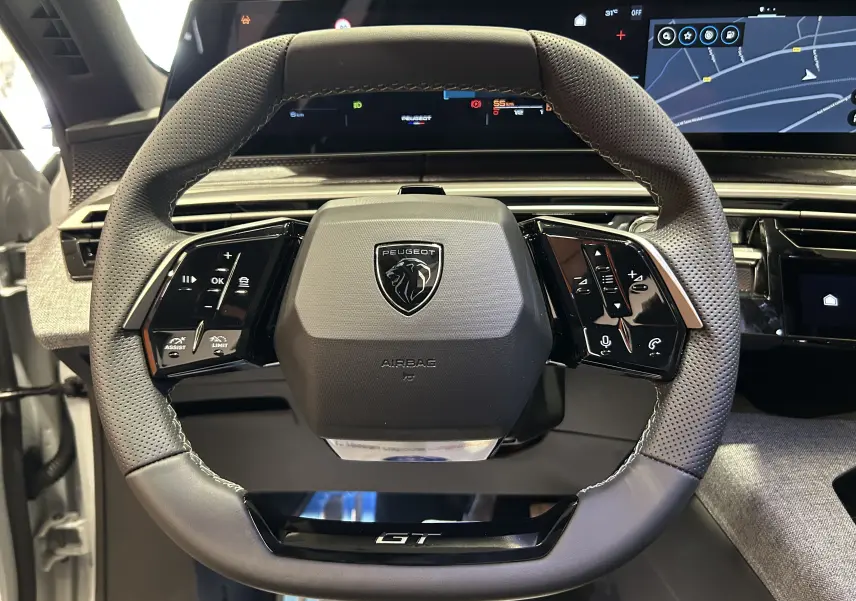 Vue rapprochée du volant GT perforé du Peugeot 3008 Hybrid 2026 avec commandes intégrées et écran numérique en arrière-plan.