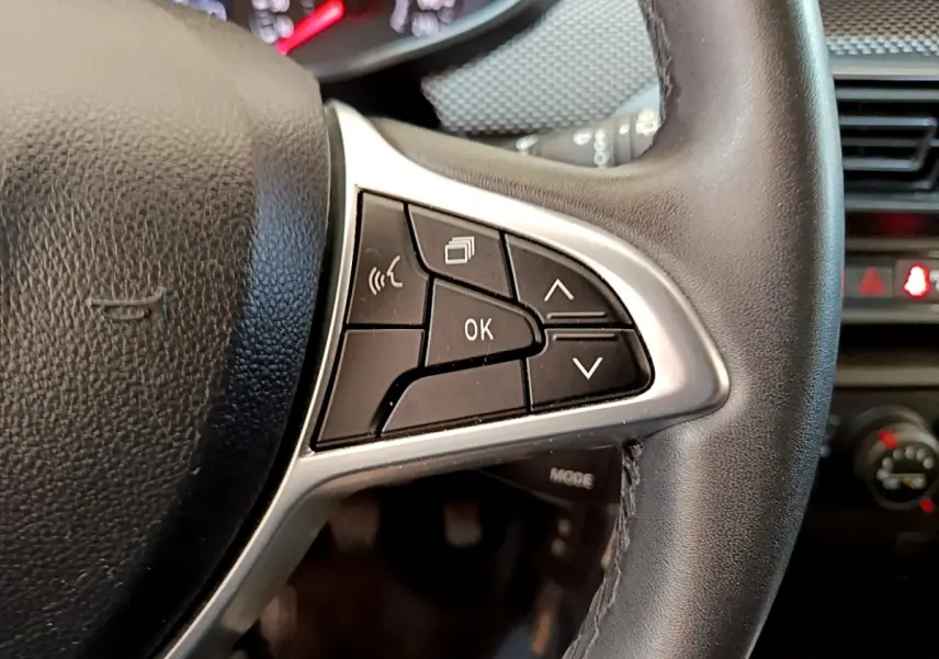 Gros plan sur les commandes multifonctions noires et argentées du volant d'une Dacia Sandero blanche, tableau de bord en arrière-plan.