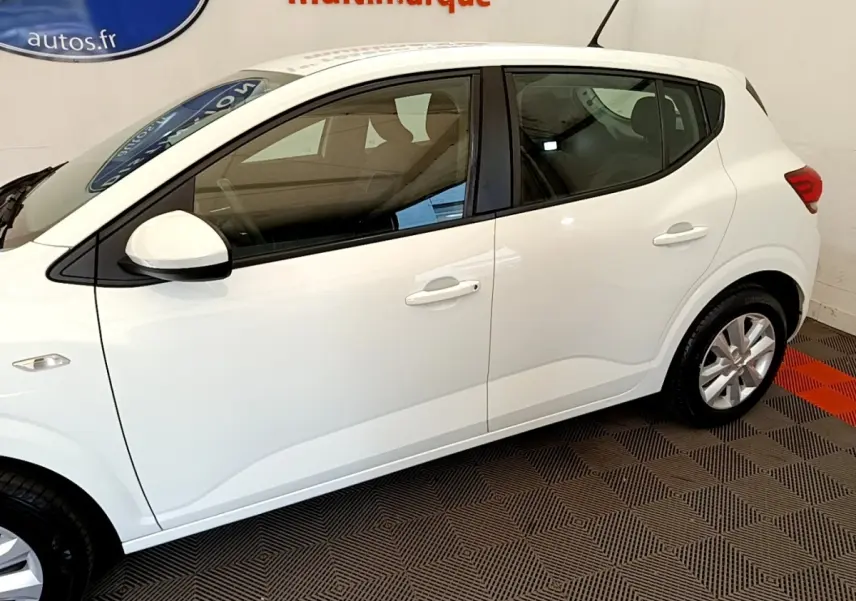Vue latérale droite d'une Dacia Sandero Confort TCe 90 blanche, en intérieur sur sol à motifs géométriques.