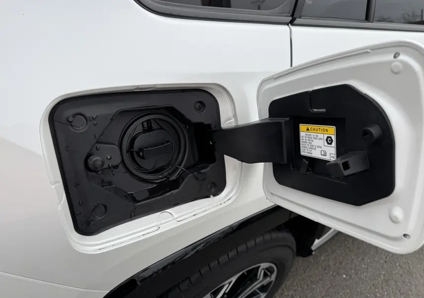 Trappe de recharge ouverte côté gauche d’un Suzuki Across blanc 2023 hybride rechargeable, détail du bouchon et étiquette de sécurité.