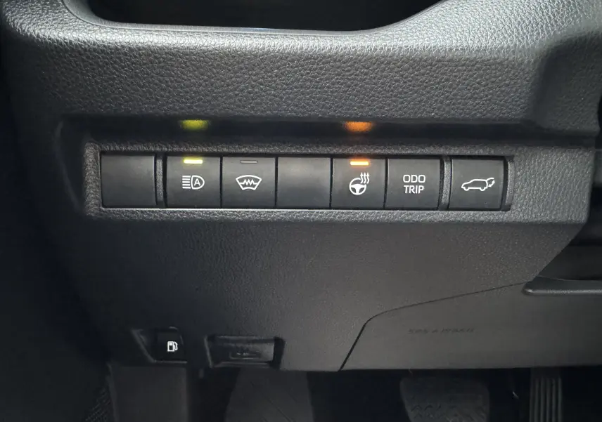 Gros plan sur les commandes du tableau de bord du Suzuki Across blanc 2023, avec voyants allumés pour phares et volant chauffant.