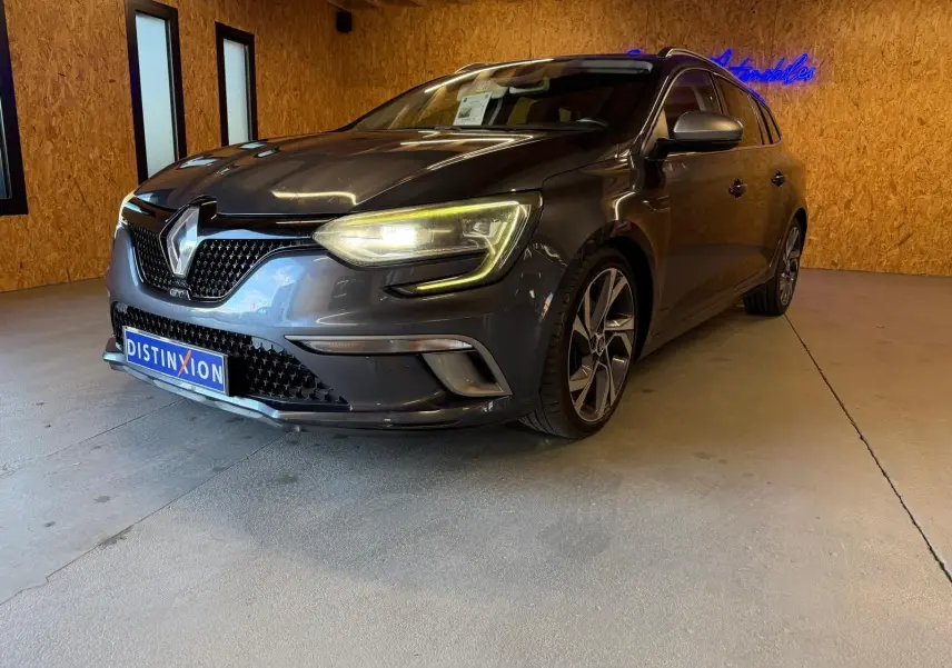 Renault Mégane Break GT gris vue 3/4 avant droit, phares allumés, dans un intérieur avec murs en bois.