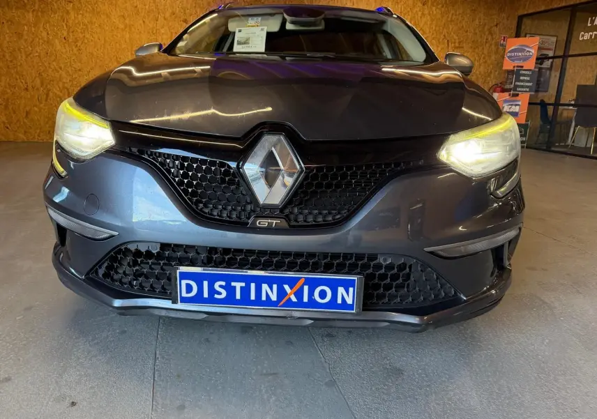 Vue de face d'une Renault Mégane Break GT gris avec phares allumés et calandre noire en intérieur showroom.