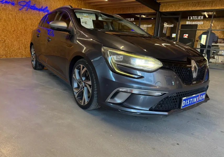 Renault Mégane Break GT gris en 3/4 avant droit, phares allumés, jantes alliage et intérieur visible en showroom.