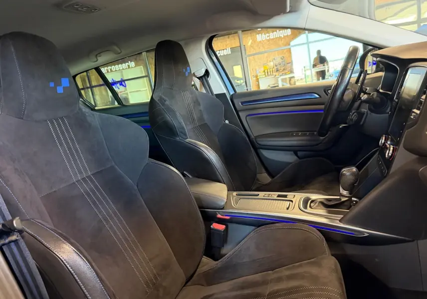 Vue intérieure côté conducteur de la Renault Mégane Break GT 2017, sièges sport noirs avec surpiqûres blanches et éclairage d'ambiance bleu.