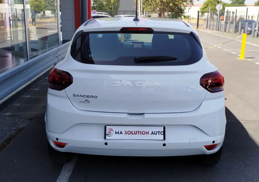 Vue arrière d'une Dacia Sandero blanche Glacier 2025 avec feux arrière foncés et logo visible sur un parking extérieur.