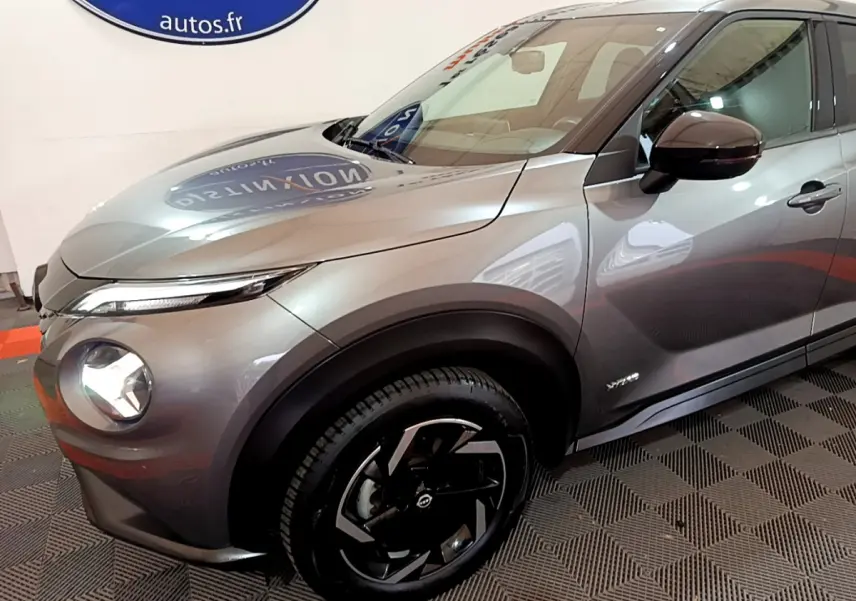 Nissan Juke Hybrid gris foncé vue 3/4 avant droit, mettant en valeur ses phares LED et jantes noires distinctives.