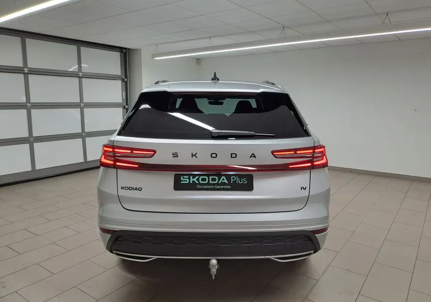Vue arrière d'un Skoda Kodiaq gris argent 2025 avec feux LED allumés et crochet d'attelage pivotant en intérieur showroom.
