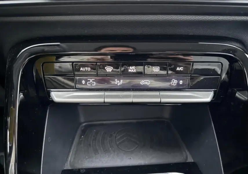 Gros plan sur la commande de climatisation automatique et le plateau de recharge sans fil noir dans une Citroën C3 gris 2025.