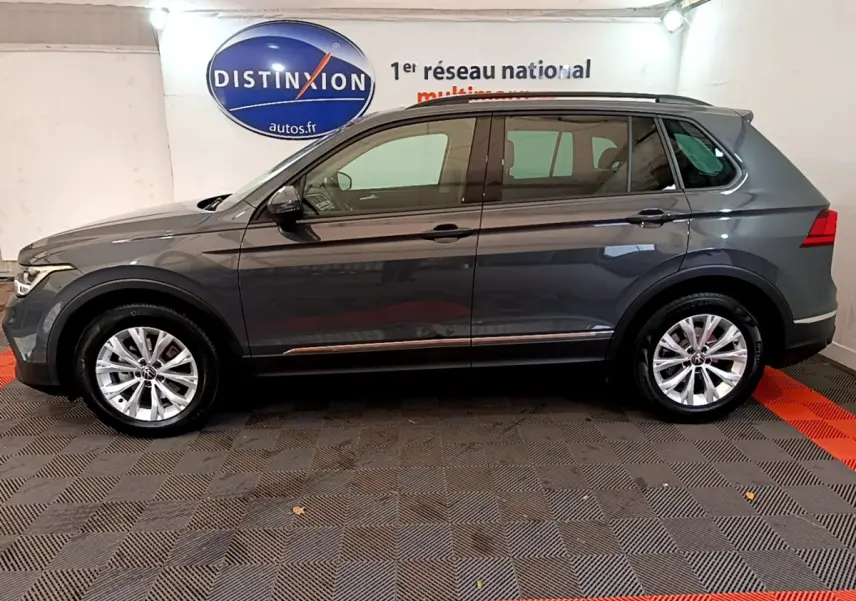 Profil côté gauche d'un Volkswagen Tiguan gris clair 2021 garé en intérieur avec jantes alliage et barres de toit noires.