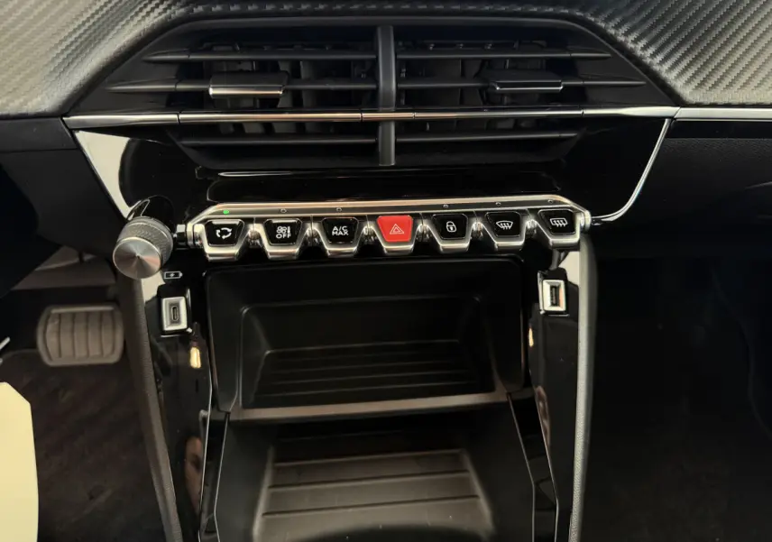 Gros plan sur la console centrale noire d'une Peugeot 208 2021, avec boutons de commande et prises USB visibles.