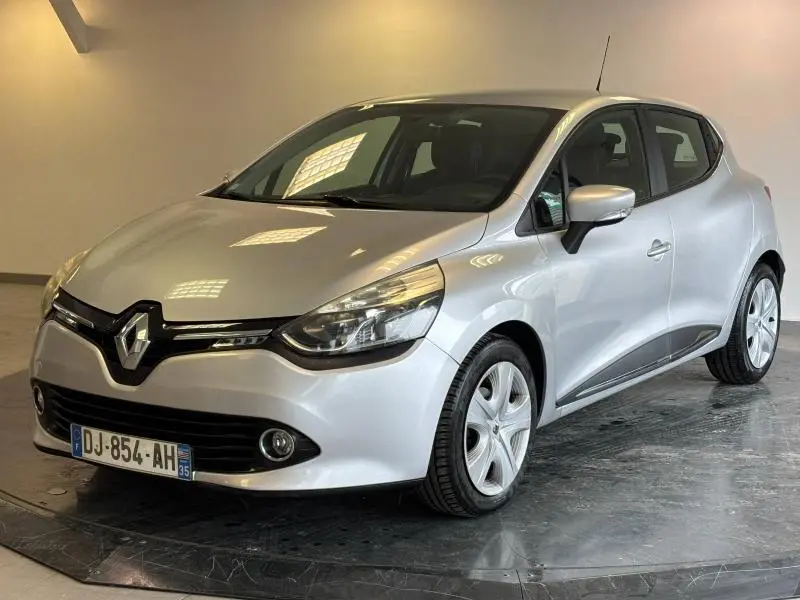 Renault Clio gris platine vue 3/4 avant droit, avec phares halogènes et jantes alu visibles en intérieur neutre.
