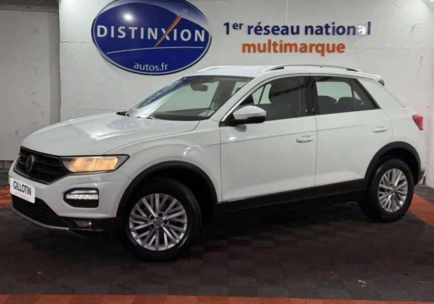 Volkswagen T-Roc blanc en vue de profil côté gauche, avec jantes alliage et barres de toit noires visibles.