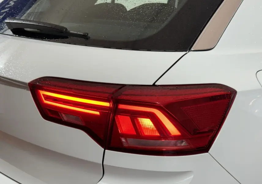 Gros plan sur le feu arrière droit rouge allumé d'un Volkswagen T-Roc blanc, avec gouttes de pluie visibles sur la vitre arrière.
