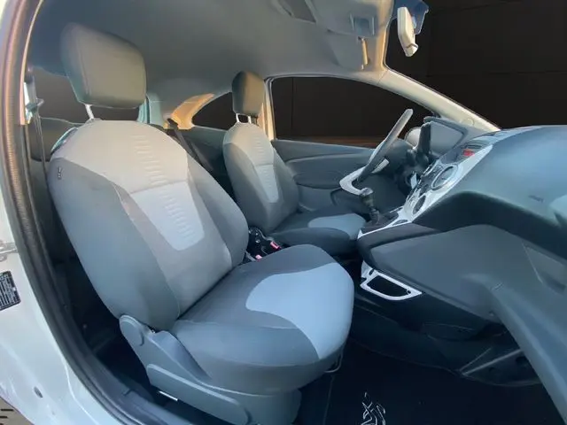 Vue intérieure côté conducteur du Ford Ka blanc 2015, montrant les sièges tissu gris et la console centrale avec boîte manuelle.