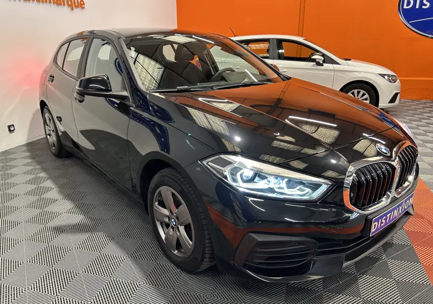 BMW Série 1 16d noire vue en 3/4 avant droit avec phares allumés dans un showroom intérieur.
