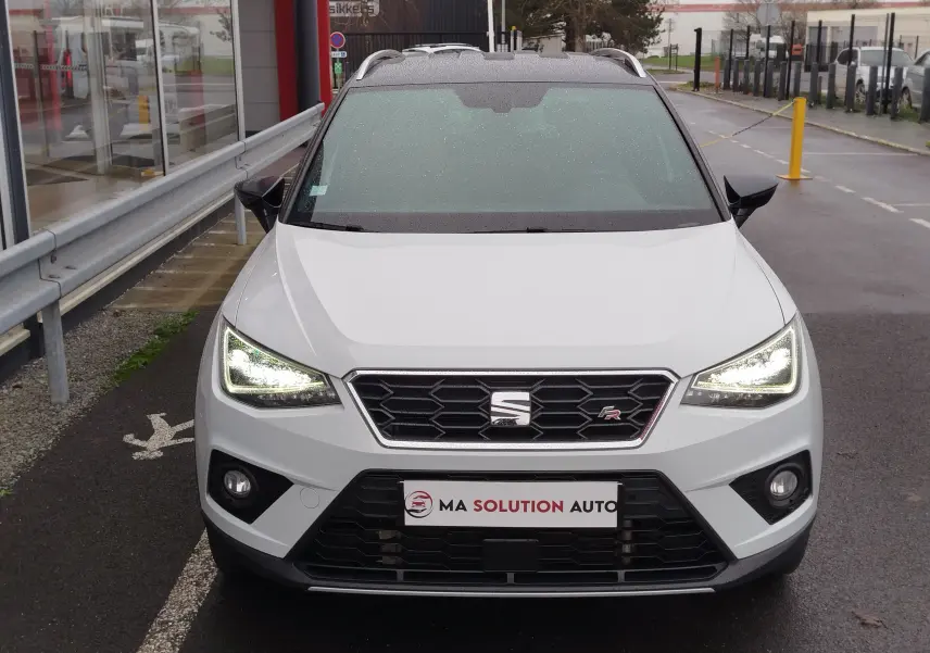 Vue avant d'un SEAT Arona blanc avec toit gris magnétique, phares allumés, garé devant un bâtiment sous la pluie.