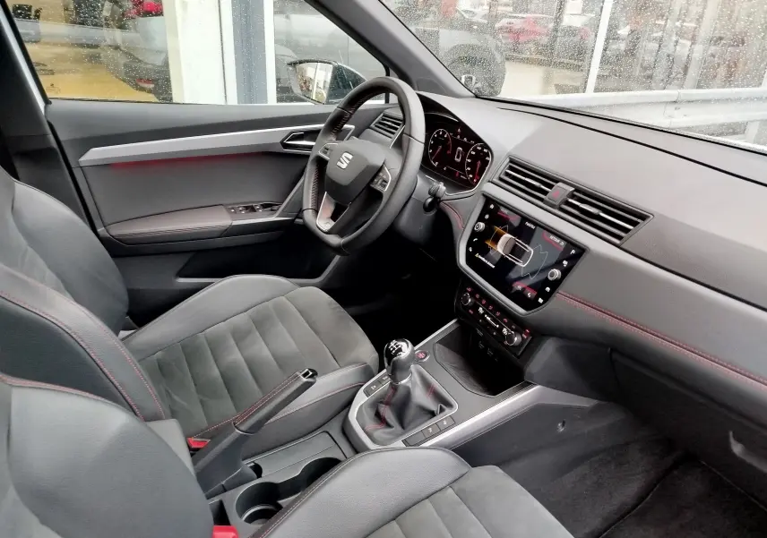 Vue intérieure côté conducteur du SEAT Arona 2020, montrant le volant, la console centrale avec boîte manuelle et les sièges en cuir noir avec surpiqûres rouges.