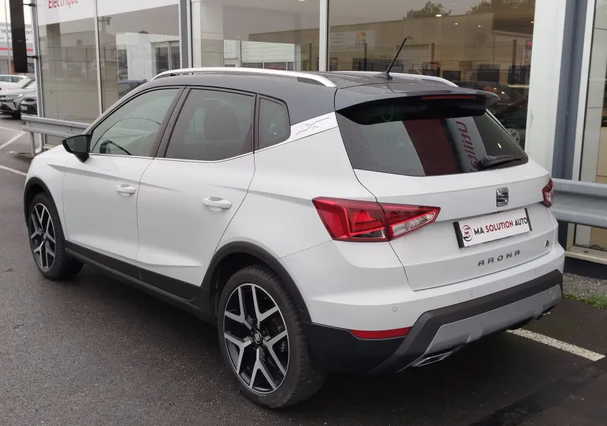 Vue 3/4 arrière droite d’un SEAT Arona blanc avec toit gris magnétique et jantes alliage noires, stationné devant un showroom.