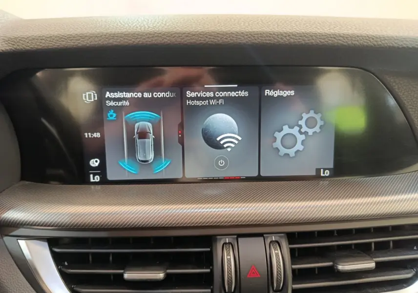 Écran tactile central affichant l'assistance à la conduite, services connectés et réglages dans un Alfa Romeo Stelvio gris foncé.