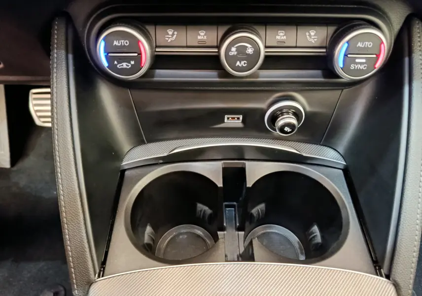 Gros plan sur la console centrale noire de l'Alfa Romeo Stelvio 2024 avec commandes climatisation et prises USB et 12V.