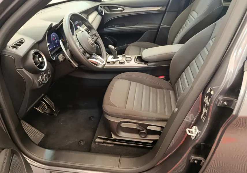 Vue intérieure côté conducteur de l'Alfa Romeo Stelvio gris foncé, avec sièges tissu et tableau de bord moderne.
