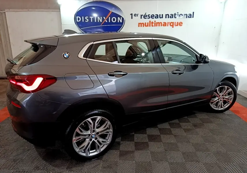 BMW X2 sDrive18i gris foncé vue de profil droit en intérieur avec feux arrière allumés et jantes alliage visibles.