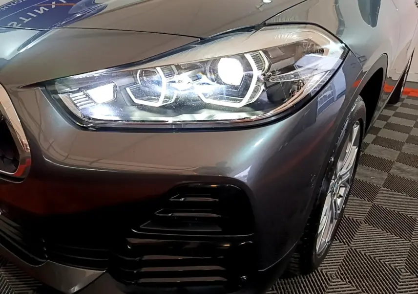 Gros plan sur le phare avant droit allumé d’un BMW X2 gris foncé, avec partie de la roue visible.